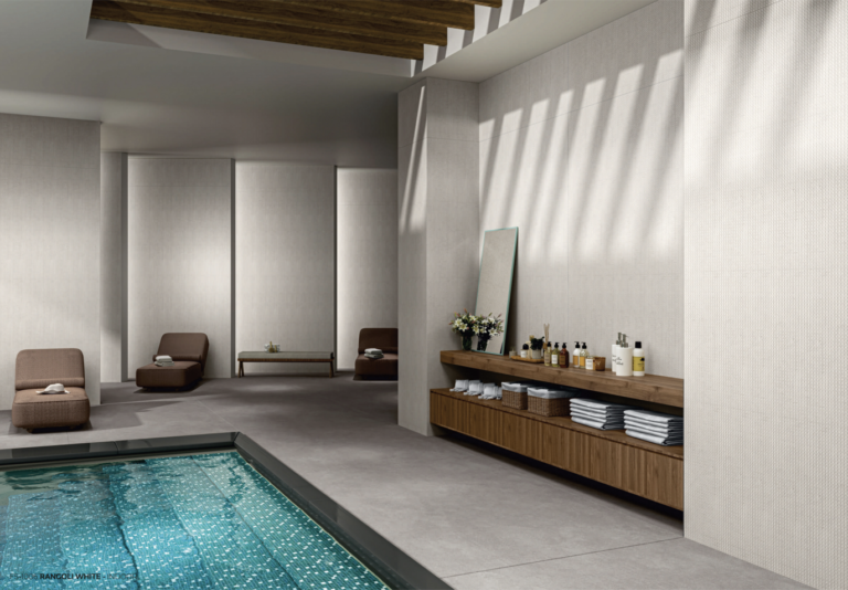 Modern indoor spa lounge featuring a tiled pool, minimalist brown lounge chairs, a wooden ceiling accent, and a sleek wooden counter with towels, toiletries, and a mirror against light-textured walls.